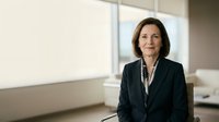 Middle-aged woman with dark hair and fair complexion wearing professional black blazer and white blouse in modern office setting with large windows and neutral background.