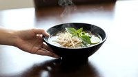 Hot steaming pho soup bowl with fresh herbs and noodles being served by hand on wooden table for Asian cuisine restaurant menu design.