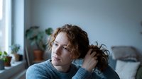 Thoughtful young Caucasian woman with curly red hair touching her hair, looking away with a pensive expression in a cozy home interior.