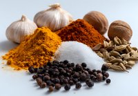 Aromatic spices and seasonings arranged on white surface including turmeric, paprika, salt, black peppercorns, garlic bulbs, nutmeg, and fennel seeds for cooking.