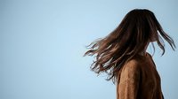 Young woman with flowing brown hair in motion against blue sky background wearing cozy sweater capturing freedom and natural movement outdoors.