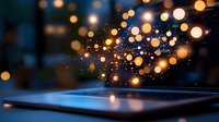 Magical glowing particles floating above digital tablet screen with bokeh light effect in dark room, creating ethereal atmosphere for technology concept.