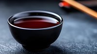 Black ceramic bowl with dark red tea on textured slate surface, minimalist Asian beverage composition with selective focus and blurred chopsticks background.