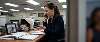 Professional businesswoman talking on phone while working at office desk with computer and documents in modern workplace environment.