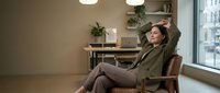 Young professional woman relaxing in modern office chair with hands behind head, taking break from work in contemporary workspace with natural light.