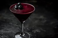 Deep purple blueberry cocktail in martini glass garnished with fresh berries against dark stone background, perfect for beverage menu photography.