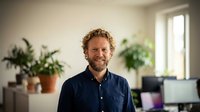 Smiling middle-aged Caucasian businessman with curly hair in navy shirt standing in modern office with plants and natural light for corporate portraits.