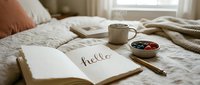 Cozy bedroom morning scene with open journal saying hello, steaming mug, fresh berries, and soft neutral linen bedding in warm light.