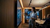 Young woman with long dark hair, wearing a light blue blazer, presenting data analytics or business strategy to colleagues in a modern conference room. She gestures toward an interactive digital display while seated professionals listen attentively during a corporate meeting or training session.