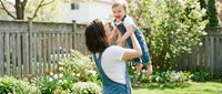 Young Caucasian mother lifting laughing toddler girl in sunny backyard garden, joyful family bonding moment outdoors in spring.
