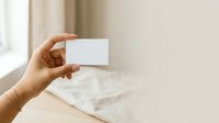 Cropped hand holding blank white business card against neutral beige interior background with soft natural light, suitable for branding and corporate mockup designs.