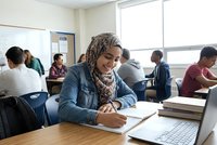 Young Middle Eastern woman in hijab studying with laptop and books in university classroom with diverse students taking exam or working on assignments.