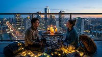 Couple enjoying romantic dinner on rooftop terrace with illuminated cityscape at twilight, featuring string lights and candles for intimate evening celebration.
