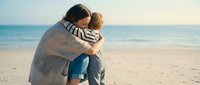 Young mother embracing toddler son on sandy beach during golden hour sunset. Tender family moment showing maternal love and bonding by ocean waves.