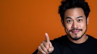 Young Asian man in black shirt pointing finger up against orange background, expressing confident and friendly attitude with warm smile.