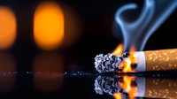 Burning cigarette with ash and smoke on reflective surface against blurred orange lights background, dramatic close-up showing addiction concept.