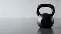 Black kettlebell weight equipment on wooden floor against white wall for strength training and fitness workout routines in minimalist gym setting.