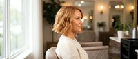 Young blonde woman with wavy bob haircut in cream sweater sitting by window in modern bright living room interior with warm lighting and contemporary decor.
