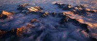 Majestic mountain peaks emerging through soft clouds at golden hour sunrise, creating dramatic alpine landscape with warm light illuminating rocky summits above misty valleys.