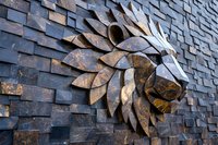 Dramatic stone wall relief sculpture featuring stylized lion head with geometric mane pattern emerging from textured brick background in dark gray and bronze tones.