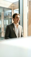 Young Caucasian male professional in charcoal blazer and white shirt gazing out modern office window with natural light, suitable for corporate, business, and leadership concepts.