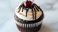 Chocolate cupcake topped with vanilla frosting, drizzled with dark chocolate sauce in spider web pattern and crowned with fresh maraschino cherry on marble surface.