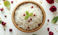 Steamed white rice in wooden bowl garnished with fresh mint leaves and dried red flower petals, surrounded by herbs and spices on white surface.