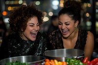 Two diverse women laughing heartily while preparing food together at night, wearing elegant party attire with bokeh lights in background, sharing joyful moment in kitchen.