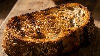 Freshly baked whole grain bread slice with seeds and nuts on rustic wooden surface, suitable for bakery, food, and culinary design projects.