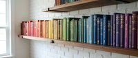Colorful books arranged by rainbow spectrum on floating wooden shelves against white brick wall creating organized home library display for reading enthusiasts.