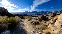 Desert hiking trail winding through rugged mountain landscape with dramatic rock formations under bright blue sky with wispy white clouds.
