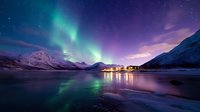 Spectacular aurora borealis dancing over snow-covered mountains and frozen lake with village lights reflecting on ice surface during polar night in Arctic landscape.