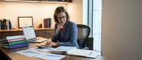 Focused Caucasian businesswoman in suit reviewing financial charts and reports at office desk with laptop, analyzing data for business strategy.