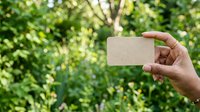 Hand holding blank kraft business card against lush green foliage background. Eco-friendly branding mockup for sustainable business, environmental marketing, or organic product promotion.