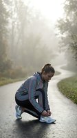 Young female athlete in colorful windbreaker adjusting shoelaces on misty tree-lined path before morning run or jog workout outdoors.
