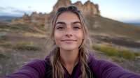 Young woman taking selfie outdoors with mountain landscape background, wearing purple sweater, natural makeup, genuine smile, casual lifestyle photo.