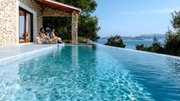 Luxury infinity pool overlooking ocean bay with stone villa and lounge chairs on terrace, crystal clear turquoise water reflecting summer sunlight.