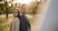 Young professional couple with glasses standing back to back in golden sunset light outdoors. Business partnership and teamwork concept for corporate marketing.