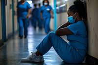 Exhausted African American female healthcare worker in scrubs and face mask sitting on hospital floor during pandemic, showing emotional strain and burnout.