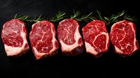 Five raw ribeye beef steaks with rich marbling arranged in a row on dark slate, garnished with fresh rosemary sprigs.