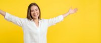 Young woman with medium-length brown hair, Caucasian ethnicity, wearing white button-up shirt with arms spread wide in celebratory gesture against bright yellow background.