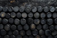 Stacked black oil barrels in industrial storage facility, showing weathered tops with metallic reflections against dark background.