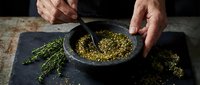 Hands grinding dried herbs with mortar and pestle on dark slate surface. Natural spice preparation for cooking and seasoning culinary ingredients.