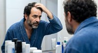 Middle-aged man with beard examining hair loss in bathroom mirror, touching forehead with concerned expression. Hair care and male grooming concept.