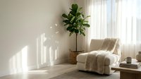 Cozy modern living room interior with comfortable armchair, fiddle leaf fig plant, and natural sunlight streaming through white curtains creating peaceful atmosphere.