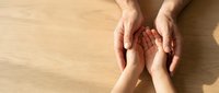 Adult hands gently holding child hands in protective caring gesture on wooden surface showing family support love and connection between generations.