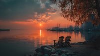 Tranquil lakeside sunset with two empty chairs on wooden dock, calm water reflecting orange sky, sailboats anchored in distance, peaceful evening atmosphere.