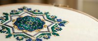 Traditional embroidery hoop with intricate geometric mandala pattern in blue and green threads on white fabric for crafting and textile art projects.