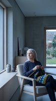 Senior woman with long gray hair, Caucasian ethnicity, sitting in modern chair by window knitting colorful yarn in minimalist concrete interior space with natural light and garden view.