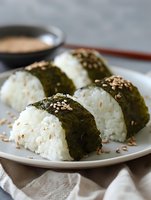 Fresh rice balls wrapped in crisp nori seaweed, topped with toasted sesame seeds on white ceramic plate. Traditional Asian cuisine with dipping sauce.
